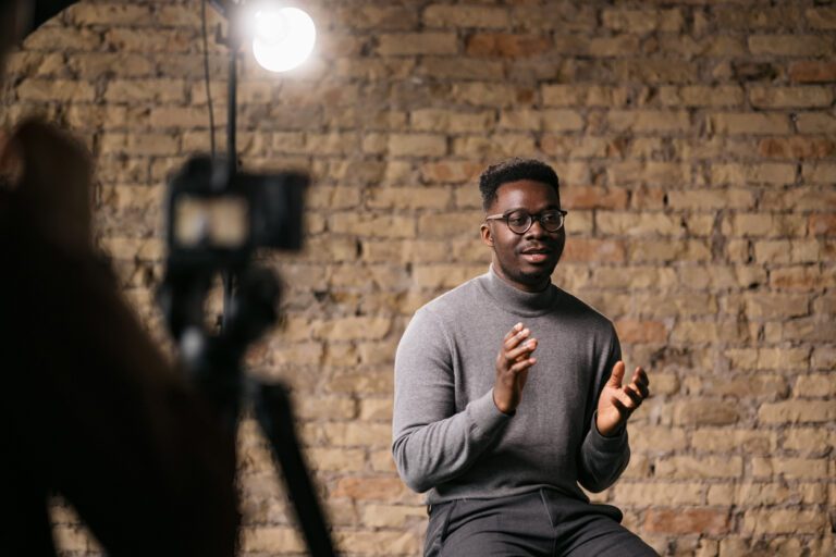 Black actor wearing glasses and a gray sweater recording a self tape in front of a brick wall, camera just out of focus.