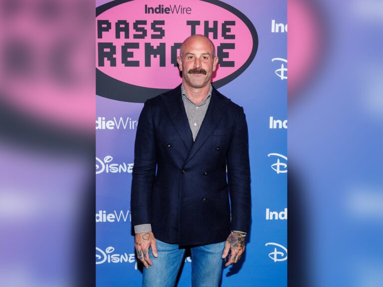 Seth Yanklewitz wearing jeans and a blazer smiling in front of a blue step-and-repeat banner.